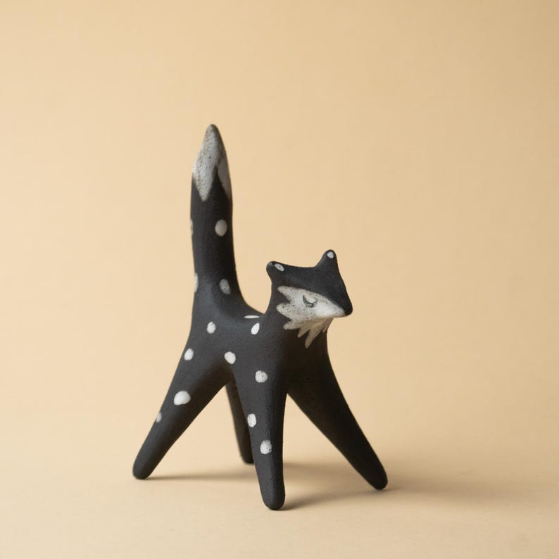 May include: A black fox ornament with white polka dots and a white-tipped tail. The fox has a stylized design with a slender body and legs. The fox's face is white with closed eyes, giving it a whimsical appearance.