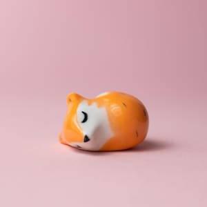 May include: A small ceramic figurine of a sleeping fox. The fox is orange and white with black eyes and a black nose. It is lying on its side with its head resting on its paws.