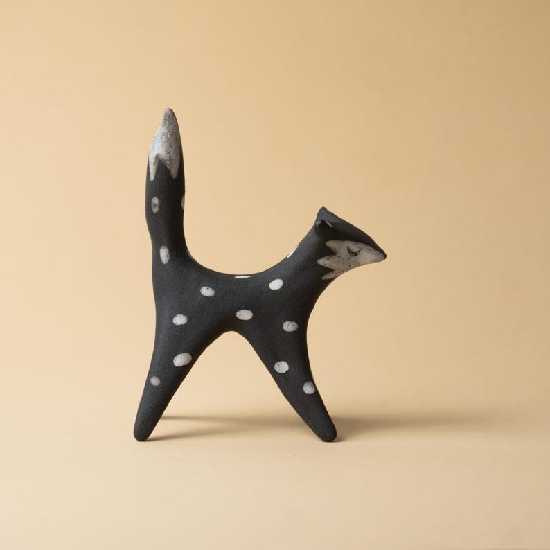May include: A black ceramic fox ornament decorated with white spots. The fox has a high, curved tail and a pointed face. The tail and face tips are a lighter shade. The ornament is set against a neutral backdrop.