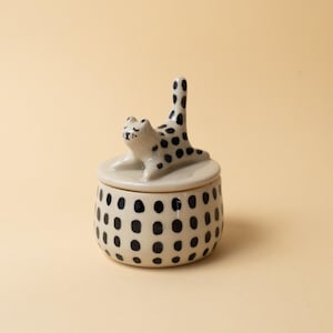 Puede incluir: Un pequeño tarro de cerámica de color crema con una tapa que presenta una figura de gato esculpida. El tarro y el gato están decorados con manchas ovaladas irregulares pintadas a mano en negro. El gato se muestra en una postura de estiramiento.