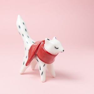 May include: A white ceramic figurine of a fox wearing a red scarf. The fox has black spots and is standing on a pink surface.