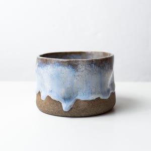 Puede incluir: Un cuenco de cerámica hecho a mano con un diseño único. El cuenco presenta una base marrón con un esmalte azul claro que gotea por los lados. El borde es de color marrón más oscuro. Este cuenco decorativo es perfecto para guardar objetos pequeños o como pieza decorativa.