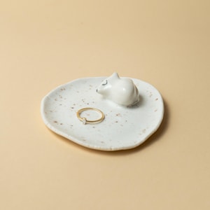 Pode incluir: Uma tigela de anel de cerâmica branca com uma pequena figura de rato a dormir. A tigela tem um design salpicado e contém um anel de ouro com uma pequena pedra transparente. A tigela de anel está sobre uma superfície bege clara.