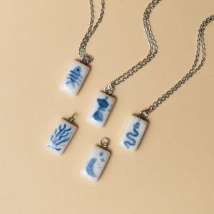 Può includere: Cinque ciondoli rettangolari in ceramica con illustrazioni blu su catene d'argento. I disegni includono uno scheletro di pesce, una caffettiera, corallo, un serpente e una luna crescente con stelle. I ciondoli sono bianchi con disegni blu.