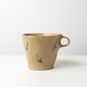Könnte beinhalten: Eine beige Keramiktasse mit rundem Henkel. Die Tasse hat eine leicht ausgestellte Form und ist mit zarten, handgemalten Zweigen in Schwarz und Orange verziert. Die Tasse eignet sich für Kaffee, Tee oder andere Getränke.