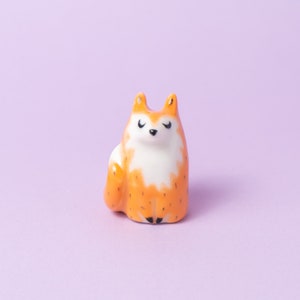 May include: A small ceramic fox figurine with orange and white fur. The fox is sitting with its paws tucked under its body and has a cute, cartoonish expression.