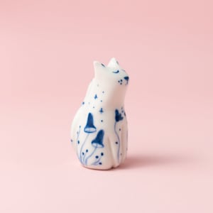 Könnte beinhalten: Eine weiße Keramik-Katzenfigur mit blauen Pilz- und Sterndesigns. Die Katze sitzt mit geschlossenen Augen. Die Figur hat eine glänzende Oberfläche und steht vor einem rosa Hintergrund.