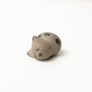 Può includere: Una piccola statuetta in ceramica color beige maculato a forma di gatto raggomitolato. Il pezzo presenta una superficie ruvida e sabbiosa con dettagli dipinti in grigio scuro che rappresentano occhi chiusi e macchie rotonde sul dorso.