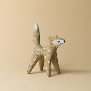 May include: A small ceramic fox figurine with white spots and a white tail tip. The figurine is standing on a light brown surface.