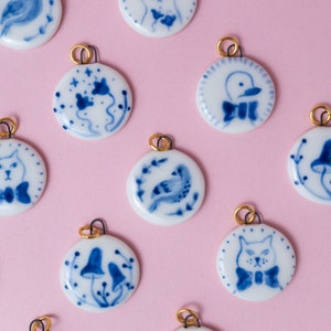 Peut inclure: Dix pendentifs en céramique avec des motifs bleus et blancs. Chaque pendentif présente un motif différent, notamment un oiseau, un chat, un champignon et une fleur. Les pendentifs sont tous enfilés sur des chaînes dorées.