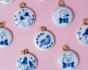 Puede incluir: Una colección de diez colgantes de cerámica con diseños azules y blancos. Cada colgante presenta un diseño diferente, incluyendo un pájaro, un gato, un hongo y una flor. Los colgantes están todos enhebrados en cadenas doradas.