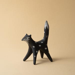 May include: A black ceramic fox figurine with white polka dots and a white-tipped tail. The fox has a stylized design with long legs and a curved back, standing on a beige surface. The fox's face is gray and white.