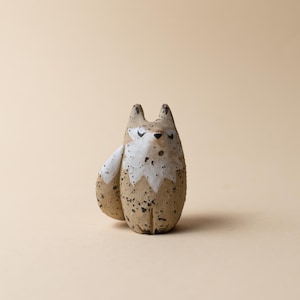 Peut inclure: Figurine de renard en céramique artisanale. Le renard a un corps beige avec des détails blancs sur la poitrine et les oreilles. La figurine a des mouchetures noires et est en position assise. Le fond est beige uni.