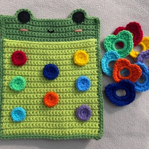 May include: A green crocheted frog-shaped pouch with a flap that opens to reveal a green felt board with colorful felt circles. The pouch is accompanied by a set of colorful crocheted rings.