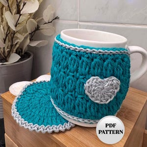 Op de afbeelding: Turquoise blauwe gehaakte mokwarmer met een grijs hartontwerp en een bijpassende turquoise blauwe gehaakte onderzetter. De mokwarmer zit op een witte mok met een grijze rand. PDF PATROON.