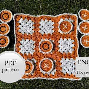 Puede incluir: Un juego de tic-tac-toe tejido a ganchillo con un esquema de color naranja y blanco. El juego incluye un tablero de juego cuadrado y piezas de juego circulares. La imagen también incluye el texto "PDF pattern" y "ENG US term".