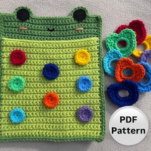 May include: Crochet frog toy with a pocket and colorful button shapes. The frog is green with black eyes and a pink mouth. The buttons are red, orange, yellow, green, blue, purple, and rainbow. The toy comes with a PDF pattern.