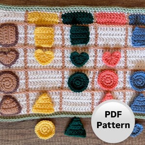 May include: A handmade crochet blanket with various shapes in shades of brown, yellow, green, coral, and blue. The blanket has a white background with a grid pattern and a green border. The image includes a "PDF Pattern" graphic.