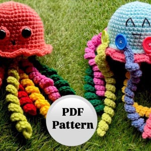 以下が含まれることがあります： 2つの編みぐるみクラゲのおもちゃ。1つは虹色の触手を持つ赤色、もう1つは紫、ピンク、黄色の触手を持つ青色。おもちゃは緑の芝生の上に置かれています。白い円の中に「PDF Pattern」というテキストがあります。