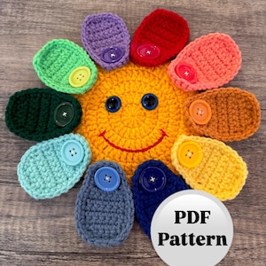 Może przedstawiać: Słoneczko wykonane na szydełku z uśmiechającą się buzią i tęczowymi płatkami. Każdy płatek ma przymocowany guzik. Słoneczko leży na drewnianej powierzchni. Wzór PDF.