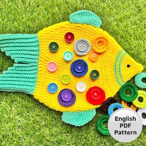 Può includere: Un pesce giallo fatto all'uncinetto con pinne e coda turchese, decorato con bottoni colorati di varie dimensioni. Il pesce è adagiato su erba verde. Un cerchio bianco con la scritta "English PDF Pattern" è nell'immagine.