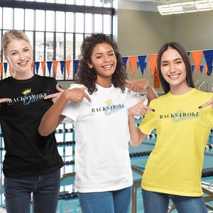 May include: Three people wearing t-shirts with the text "Backstroke Queen" and a crown graphic. The shirts are black, white, and yellow. The people are standing in front of a swimming pool.
