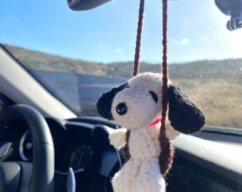Op de afbeelding: Een witte en zwarte gehaakte hond die hangt aan de achteruitkijkspiegel van een auto. De hond houdt een rood touwtje in zijn bek.