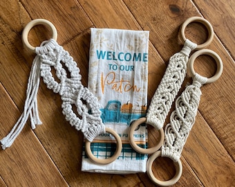 Può includere: Quattro portasciugamani in macramè con anelli in legno e un canovaccio decorativo. L'asciugamano è bianco con un camion blu e zucche, e la scritta "WELCOME TO OUR PATCH". Il macramè è bianco sporco.
