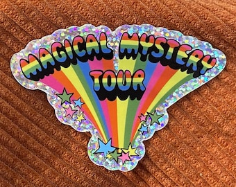 Pode incluir: Um autocolante holográfico com o texto MAGICAL MYSTERY TOUR em letras pretas, a negrito e onduladas. Um padrão de raios de sol com as cores do arco-íris irradia do centro, acentuado por pequenas estrelas multicoloridas em rosa, azul, verde e amarelo.
