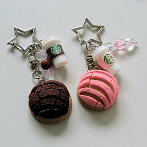 May include: Two keychains featuring a pink and brown concha pastry with a miniature Starbucks coffee cup charm. The charms are attached to a silver star-shaped keychain.