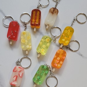 May include: Assortment of nine colorful popsicle keychains. Each keychain features a different fruit design, including watermelon, lemon, lime, orange, and strawberry. The keychains are made of clear resin with a wooden stick and a silver metal ring.