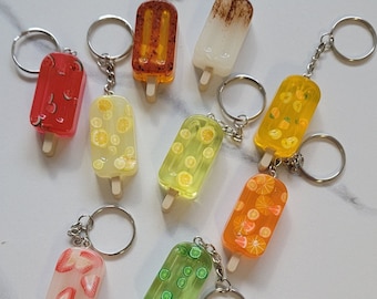 May include: Assortment of nine colorful popsicle keychains. Each keychain features a different fruit design, including watermelon, lemon, lime, orange, and strawberry. The keychains are made of clear resin with a wooden stick and a silver metal ring.