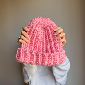 Puede incluir: Un gorro de punto grueso de color rosa brillante. El gorro está hecho de hilo grueso con un diseño acanalado y un borde doblado. El gorro se sostiene frente a una persona.