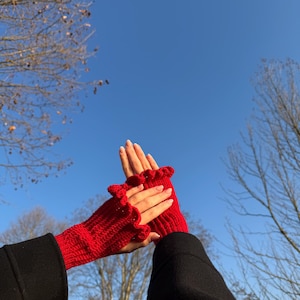 May include: A pair of bright red, knitted fingerless gloves with a ruffled edge. The gloves are held up against a clear blue sky, showcasing their vibrant color and textured design. The gloves are a stylish accessory.