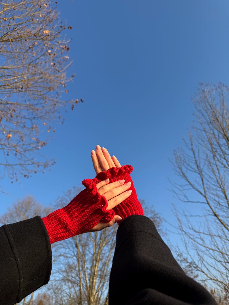 May include: A pair of bright red, knitted fingerless gloves with a ruffled edge. The gloves are held up against a clear blue sky, showcasing their vibrant color and textured design. The gloves are a stylish accessory.