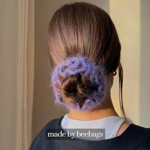 Könnte beinhalten: Ein lila-brauner gehäkelter Haarknoten-Wrap, der im Nacken getragen wird. Das Haaraccessoire ist aus weichem, flauschigem Garn gefertigt. Der Text "made by beebags" ist unten im Bild zu sehen.