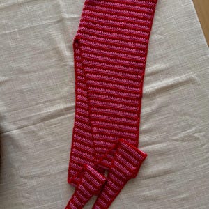 Puede incluir: Un conjunto de bufanda y calentadores de piernas a juego hechos a mano con ganchillo. El conjunto es rojo con rayas horizontales rosas. La bufanda es larga y rectangular, y los calentadores son tubulares.
