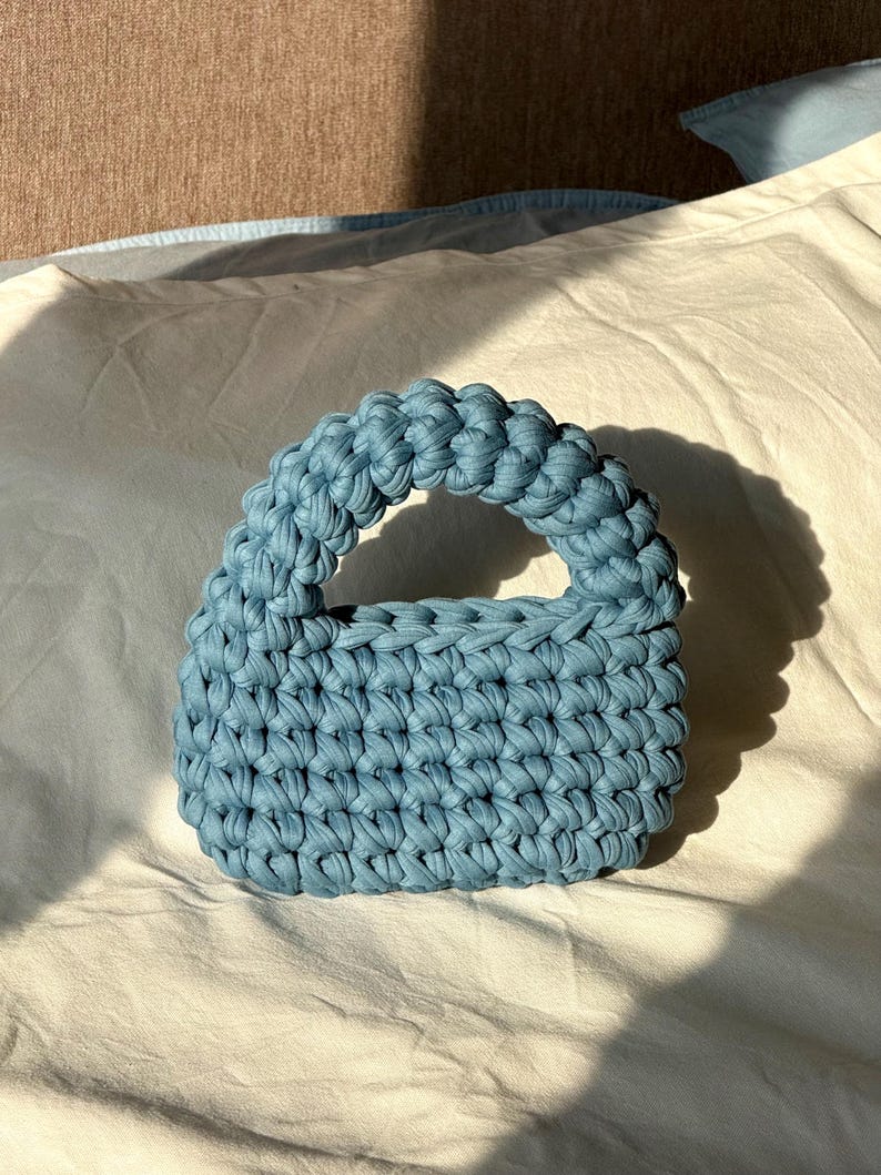 Może przedstawiać: Mała, błękitna torebka szydełkowa z zaokrąglonym uchwytem. Torba wykonana jest z grubej, teksturowanej przędzy, a ściegi są dobrze widoczne. Torba leży na białej powierzchni, z cieniami światła słonecznego.