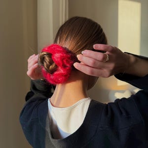 May include: A red, fluffy scrunchie wrapped around a bun hairstyle. The scrunchie is a vibrant red color and appears to be made of a soft, textured material. The person is wearing a black sweater over a white shirt.