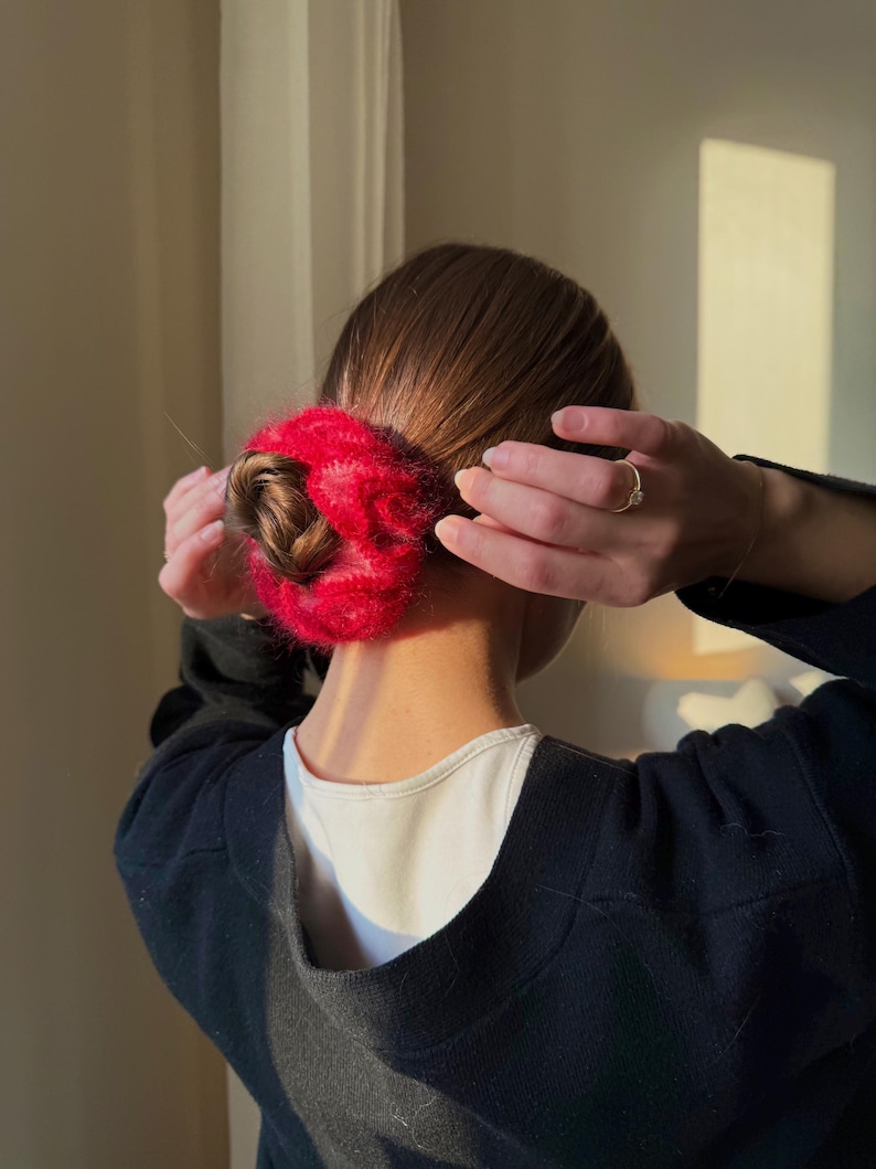 May include: A red, fluffy scrunchie wrapped around a bun hairstyle. The scrunchie is a vibrant red color and appears to be made of a soft, textured material. The person is wearing a black sweater over a white shirt.