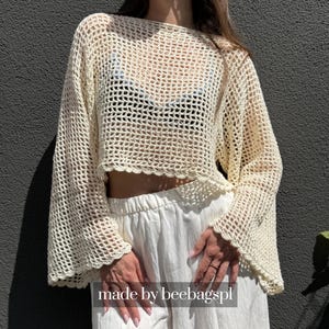 May include: A cream-colored, long-sleeved crochet top featuring an open-knit grid pattern, a wide boat neckline, and scalloped edges on the cuffs and hem. The garment is worn over a black undergarment and white pants. Text reads: made by beebagspl.