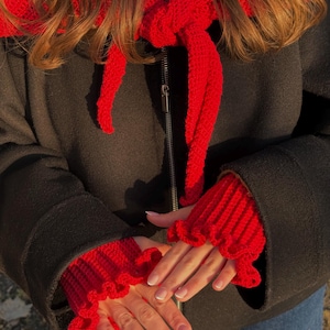 May include: A red knitted scarf and matching fingerless gloves with ruffled edges. The scarf is tied in a bow, and the gloves are worn over a black coat. The image is taken in natural light.