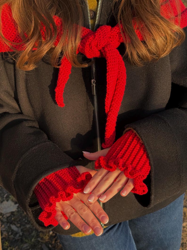May include: A red knitted scarf and matching fingerless gloves with ruffled edges. The scarf is tied in a bow, and the gloves are worn over a black coat. The image is taken in natural light.