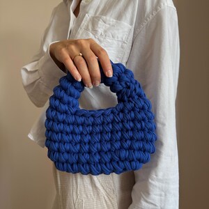 Może przedstawiać: Żywa, niebieska, szydełkowa torebka z zaokrąglonym uchwytem. Tekstura torby jest wyraźnie zdefiniowana, ukazując ciasny splot tkaniny. Torebka jest trzymana na neutralnym tle, podkreślając jej odważny kolor i unikalny design.