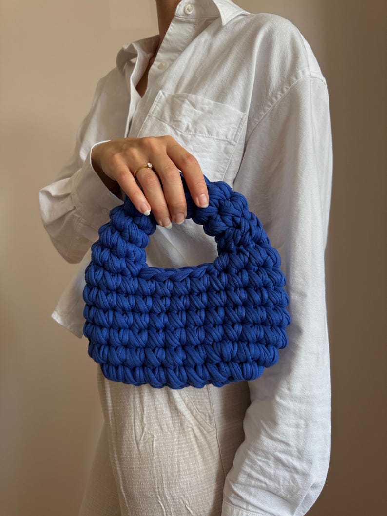 Może przedstawiać: Żywa, niebieska, szydełkowa torebka z zaokrąglonym uchwytem. Tekstura torby jest wyraźnie zdefiniowana, ukazując ciasny splot tkaniny. Torebka jest trzymana na neutralnym tle, podkreślając jej odważny kolor i unikalny design.