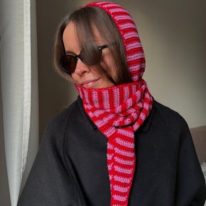 Puede incluir: Un conjunto de capucha y bufanda de crochet a rayas rojas y rosas. La capucha cubre la cabeza y el cuello, con la bufanda drapeada alrededor del cuello. El conjunto est&aacute; hecho de un hilo suave y texturizado. La persona lleva gafas de sol negras y un abrigo negro.