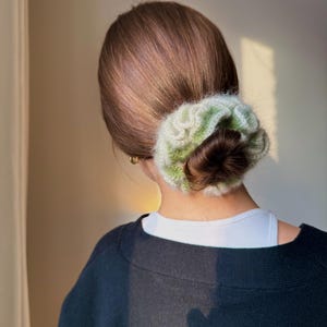 May include: A woman's hair is styled in a low bun, secured with a green and white crocheted scrunchie. She is wearing a black sweater over a white top. The background is a neutral color.