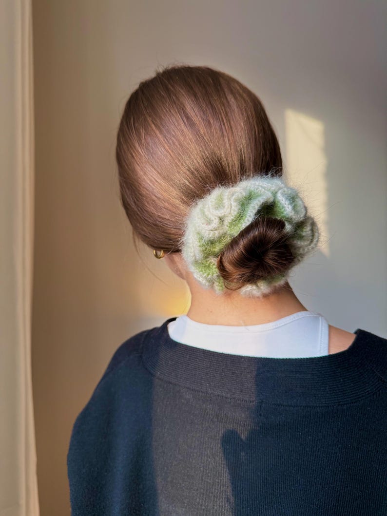 May include: A woman's hair is styled in a low bun, secured with a green and white crocheted scrunchie. She is wearing a black sweater over a white top. The background is a neutral color.