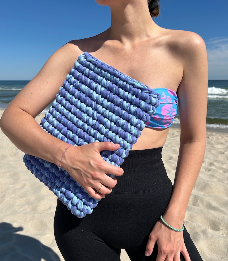 May include: A blue and light blue crocheted clutch bag with a striped pattern. The bag is held by a person wearing a blue and pink floral print bandeau top and black leggings.