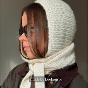 Peut inclure: Une cagoule en crochet de couleur crème avec de longs liens qui s'enroulent autour du cou. La texture du tricot est uniforme sur toute la coiffe. Le texte "made by beebagspl" est visible au centre inférieur de l'image.